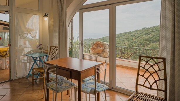 A dining area with a table and chairs, a view of a mountainous landscape through large windows, and a balcony with a rustic outdoor setting. - APP.TI STRACCOLIGNO - CASA MIMMA (Capoliveri)
