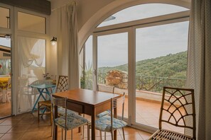 A dining area with a table and chairs, a view of a mountainous landscape through large windows, and a balcony with a rustic outdoor setting. - APP.TI STRACCOLIGNO - CASA MIMMA (Capoliveri)