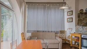 A cozy living room with a wooden dining table, beige sofa, and wicker chairs. A large window with patterned curtains allows natural light. A kitchen area with a stove and shelves is visible to the right. - APP.TI STRACCOLIGNO - CASA MIMMA (Capoliveri)