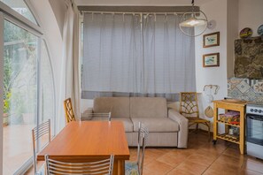 A cozy living room with a wooden dining table, beige sofa, and wicker chairs. A large window with patterned curtains allows natural light. A kitchen area with a stove and shelves is visible to the right. - APP.TI STRACCOLIGNO - CASA MIMMA (Capoliveri)