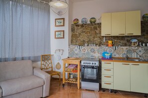 A compact living space with a sofa, a dining table, a small kitchen area with a stove, and a stone wall backsplash. - APP.TI STRACCOLIGNO - CASA MIMMA (Capoliveri)