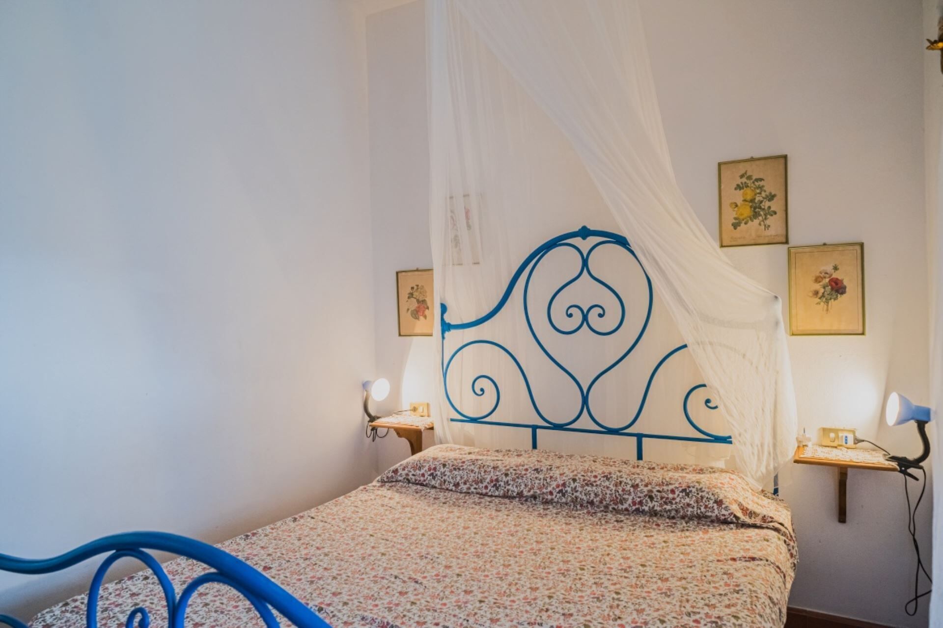 A bed with a floral bedspread, a blue metal headboard, and a white canopy. There are two wall-mounted lamps and framed floral pictures on the wall.
