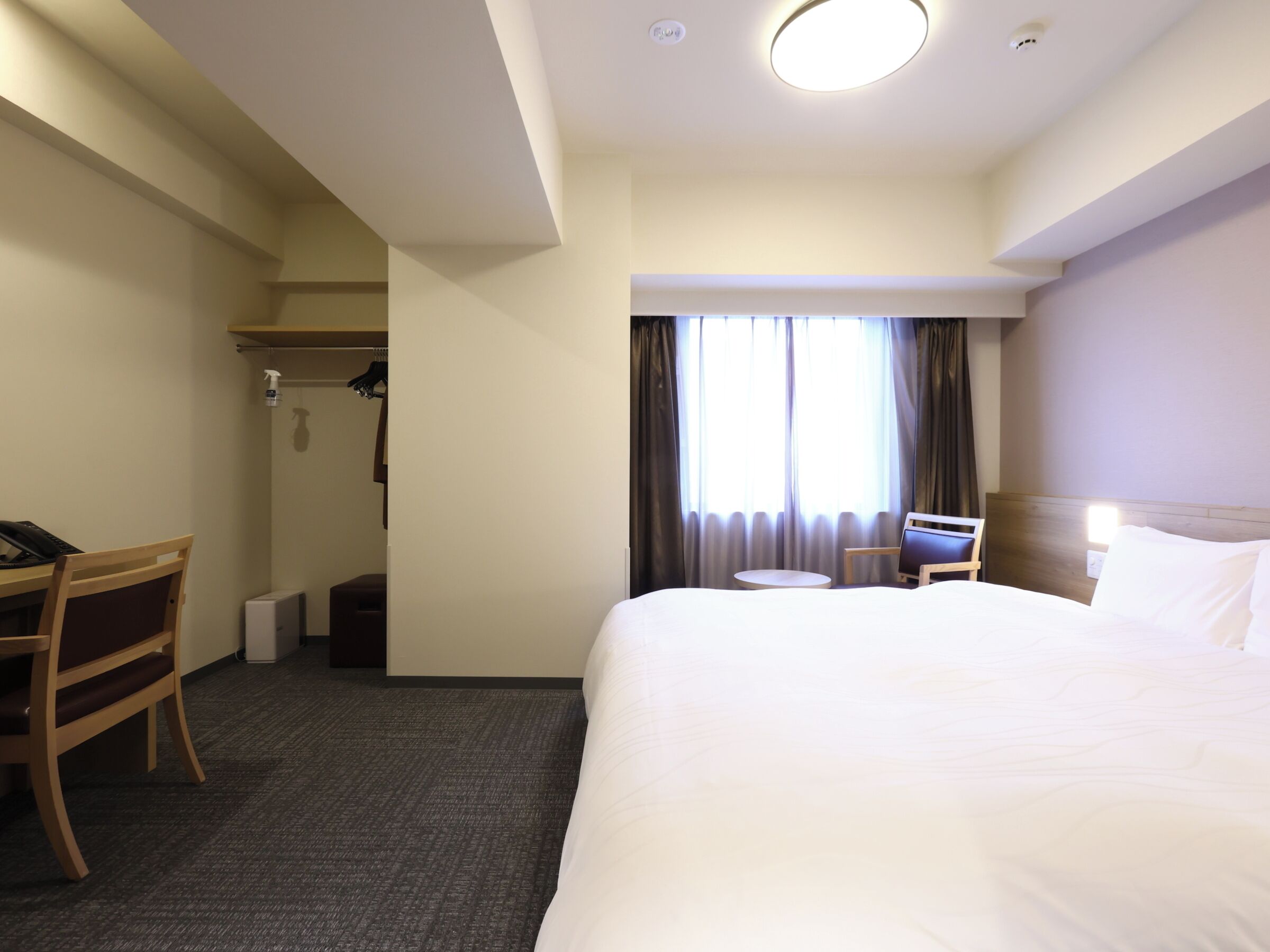 Quarto de hotel com uma cama grande, uma mesa com cadeira, uma pequena mesa redonda e um guarda-roupa.