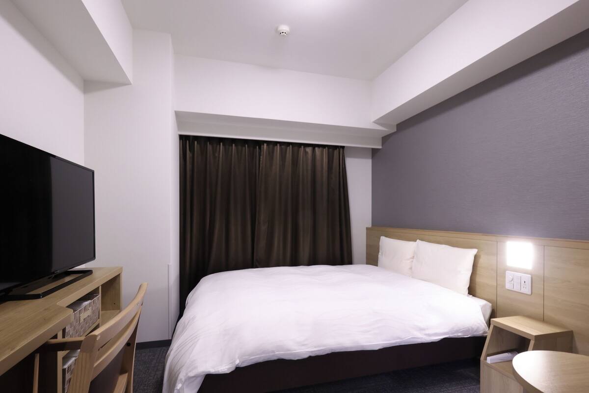Habitación de hotel con cama, televisor sobre un mueble de madera, mesita de noche y lámpara fijada a la pared.
