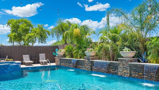 Une piscine d’un bleu éclatant, agrémentée d’une cascade en pierre, entourée d’une végétation luxuriante et d’une clôture en bois.