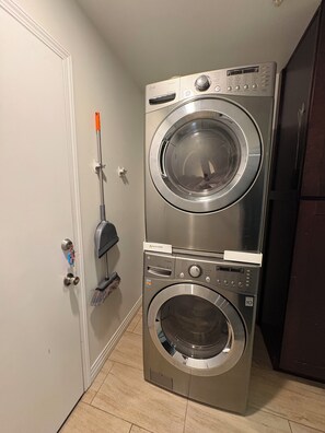 Una lavadora y secadora apiladas en un cuarto de lavado, con una puerta y una escoba apoyada en la pared.