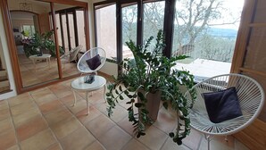 A spacious room with large windows, a round table, two white wicker chairs, and a potted plant. - Magnificent, peaceful villa surrounded by olive trees. Exceptional panoramic view (Fayence)