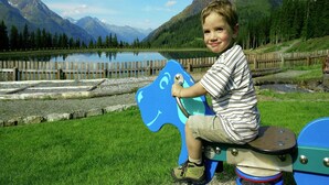 A child on a blue and white ride-on toy in a grassy area with a lake and mountains in the background. - Apartment in See near Ski Slopes (See)