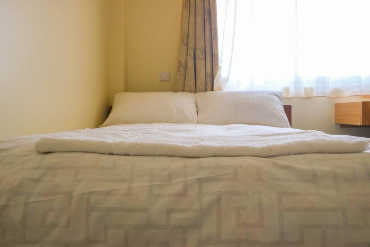A bed with a patterned bedspread, a white blanket, and a wooden nightstand with a lamp.