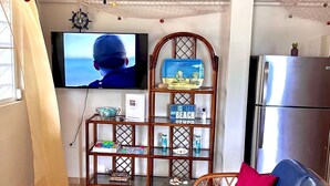 A living room with a TV, a wooden shelf with decorative items, a wicker sofa, and a refrigerator. - Beautiful, spacious house 9 minute drive to beach with electric generator (Rincón)
