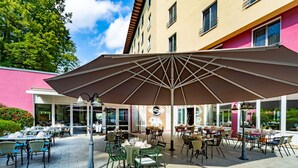 An outdoor dining area with tables and chairs, a large umbrella, and a building with a pink facade in the background. - Parkhotel DZ Standard - Parkhotel Görlitzs (Görlitz)