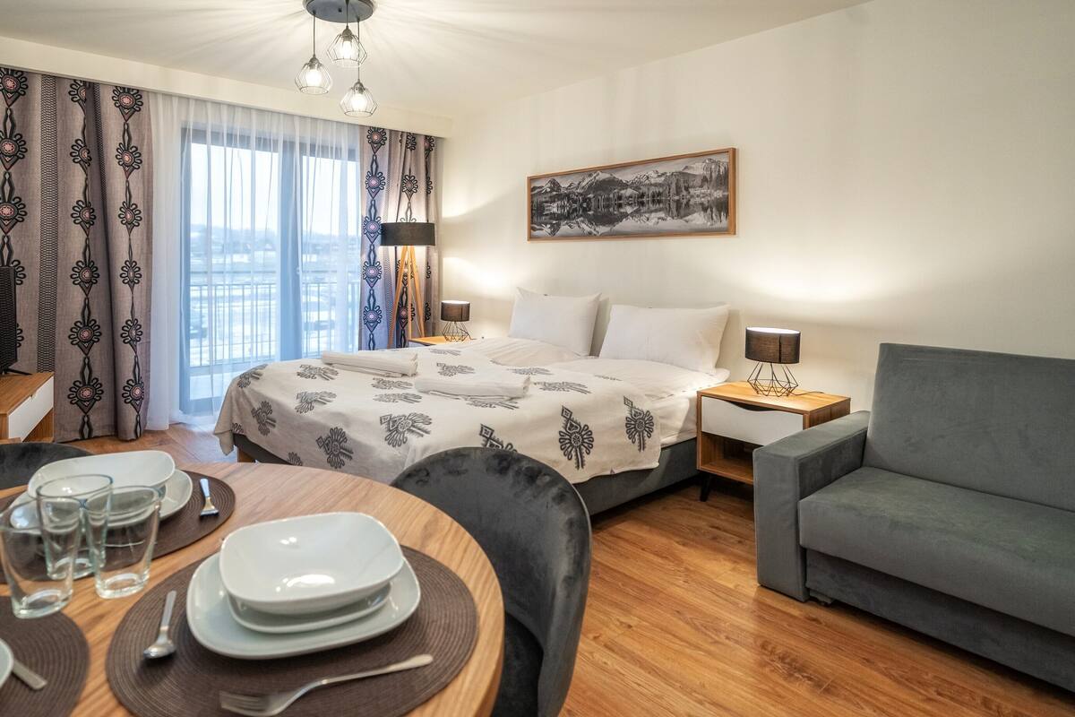 Et hotelværelse med seng, sofa, spisebord dækket med tallerkener og glas, et billede på væggen og et vindue med gardiner.