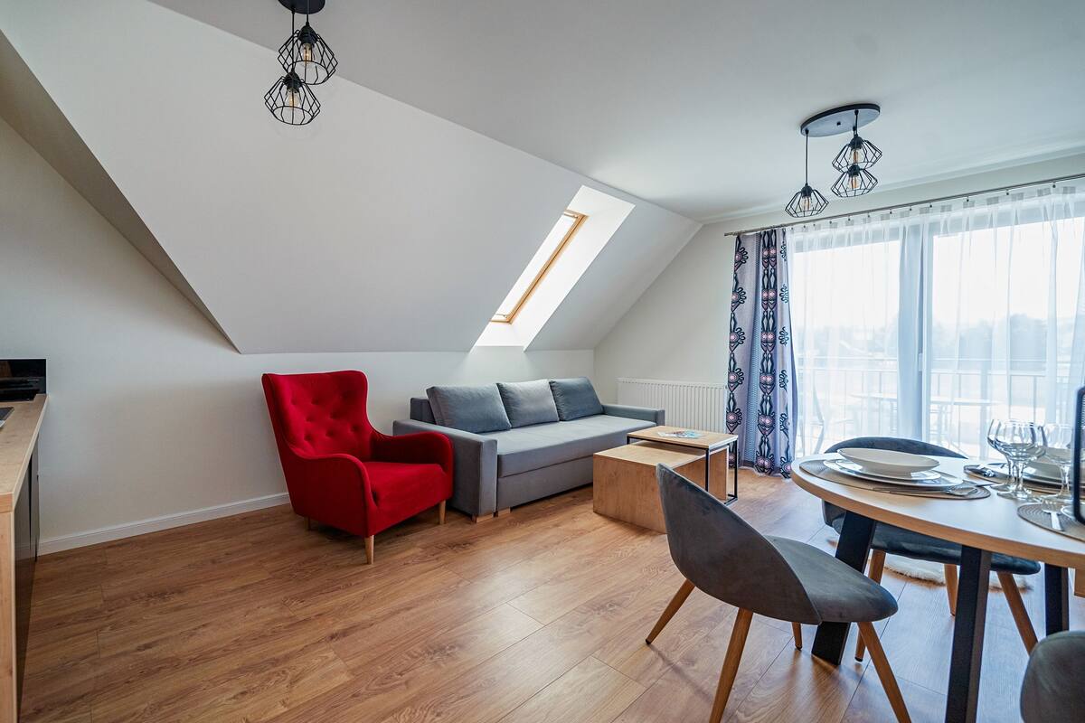 En moderne loftlejlighed med et køkken, en rød lænestol, en grå sofa, et spiseområde med et bord dækket til to personer, og et stort ovenlys.