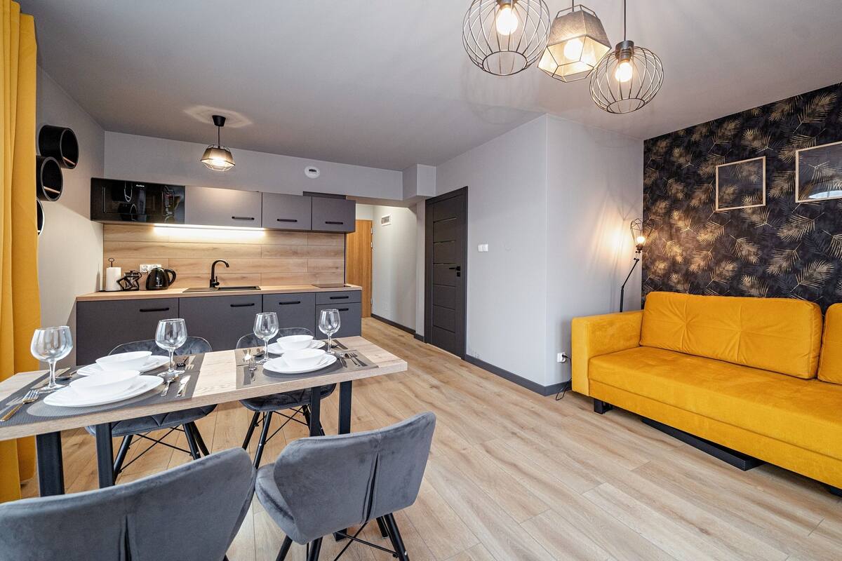 En moderne stue med en gul sofa, et spiseområde med et bord dækket til fire personer, og et køkken med mørke skabe og en mikrobølgeovn.