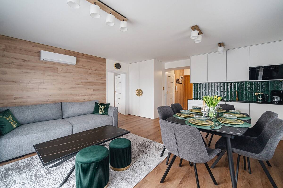 En moderne stue med en grå sofa, et spisebord dækket til fire personer, og et køkken med en grøn bagvæg.