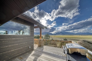 A wooden deck with a barbecue grill, a stone wall, and a view of mountains and a field.