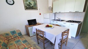 A compact kitchen with white cabinets, a stove, and a sink. There is a dining table with a white tablecloth and two chairs. A laptop is on a small table beside the sofa. - Holiday home in Rosolina Mare (Rosolina Mare (RO))