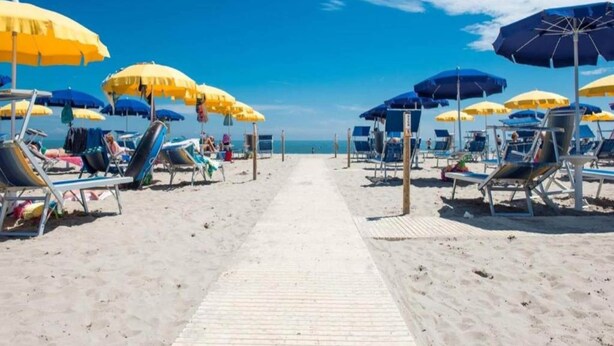 A beach with rows of sun loungers and umbrellas, a wooden walkway, and a clear view of the ocean. - Holiday home in Rosolina Mare (Rosolina Mare (RO))