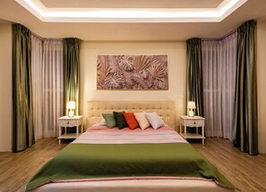 A hotel room with a large bed, two bedside tables with lamps, a ceiling light, and a tropical leaf-patterned wall art.