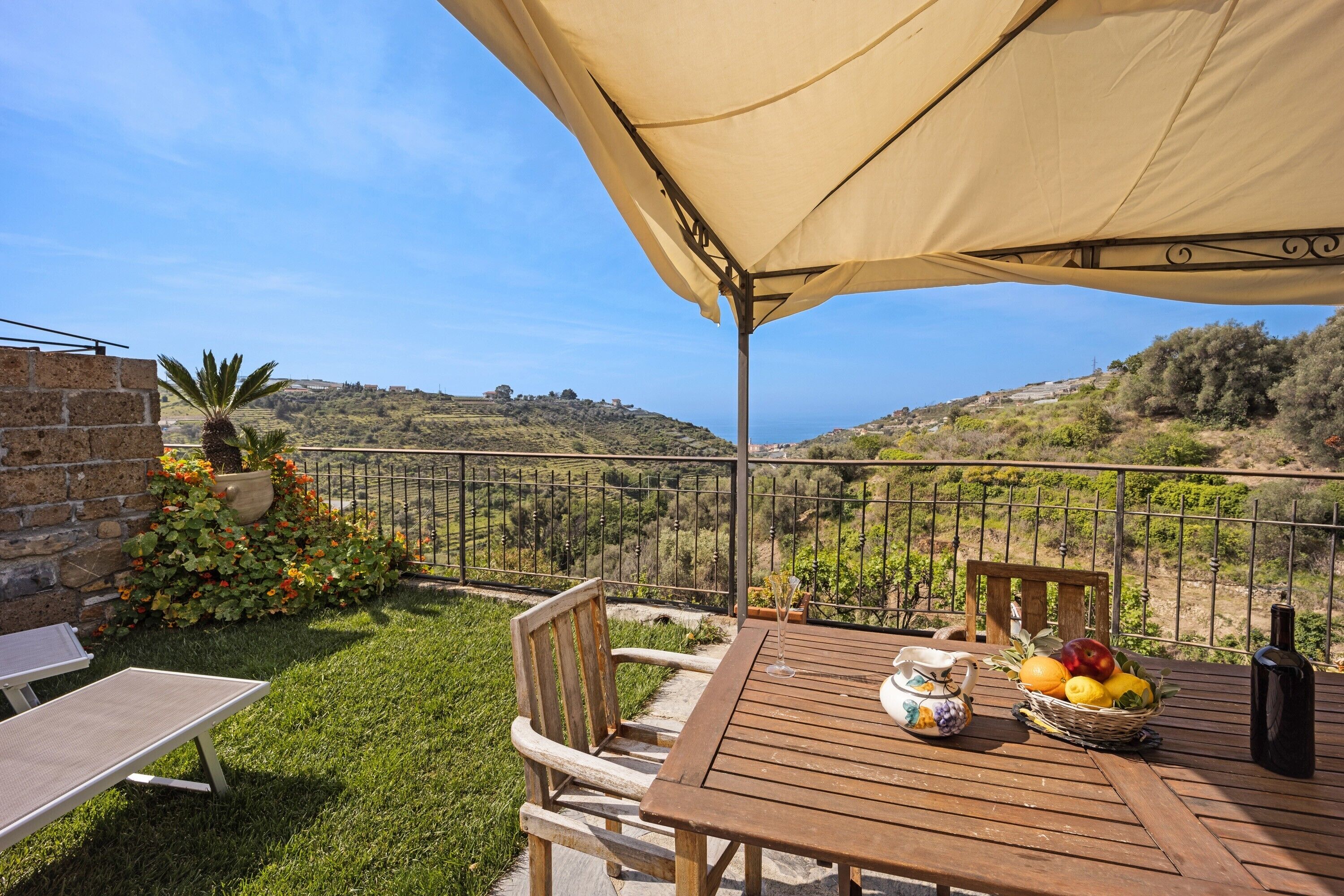 A wooden table with a wicker basket of fruit, a bottle, and a cup, set under a large canopy on a terrace with a view of hills.