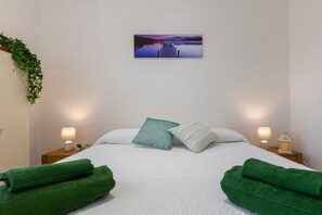 A neatly made bed with green towels, two bedside lamps, a nightstand with a plant, and a wall-mounted picture of a lakeside scene.