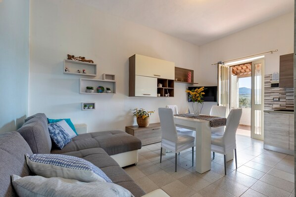 A modern living room with a sofa, dining table, chairs, and a kitchen area with shelves and a view.
