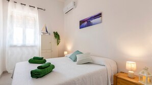Ein ordentlich bezogenes Bett mit grünen Handtüchern, eine Nachttischlampe, ein Nachttisch, eine wandmontierte Klimaanlage, ein gerahmtes Bild und ein Fenster mit transparenten Vorhängen.