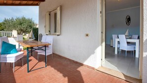 Eine überdachte Terrasse mit Tisch und Stühlen, eine Tür zu einem Essbereich im Innenbereich und Blick auf Bäume.