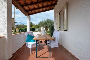 A covered patio with a wooden table and two white chairs, a white wall, and a view of greenery.