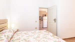 A bedroom with a bed, a nightstand, a lamp, and a door leading to a kitchen.
