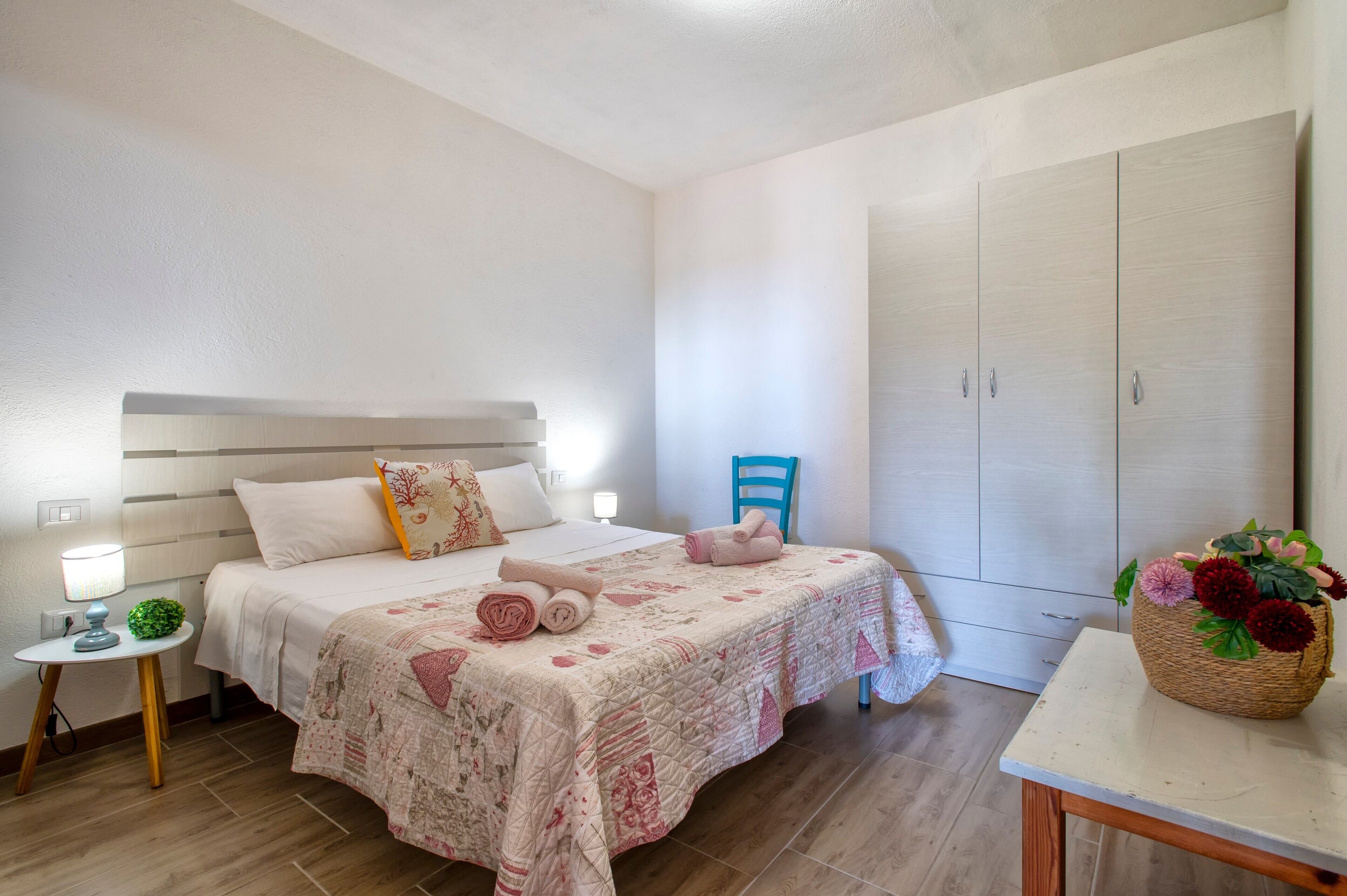A bedroom with a bed, bedside table, lamp, chair, wardrobe, and a decorative basket.