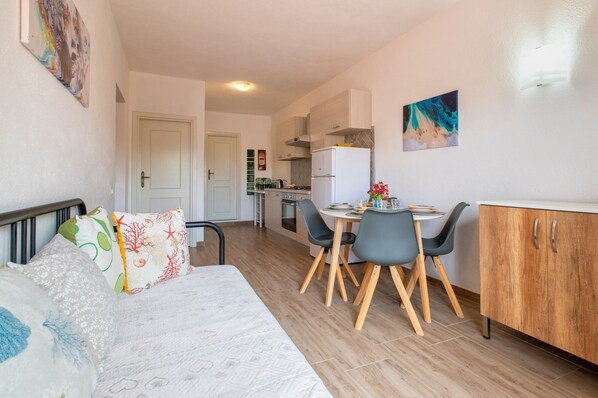 A compact living space with a bed, a dining table with chairs, a kitchenette, and a small cabinet.