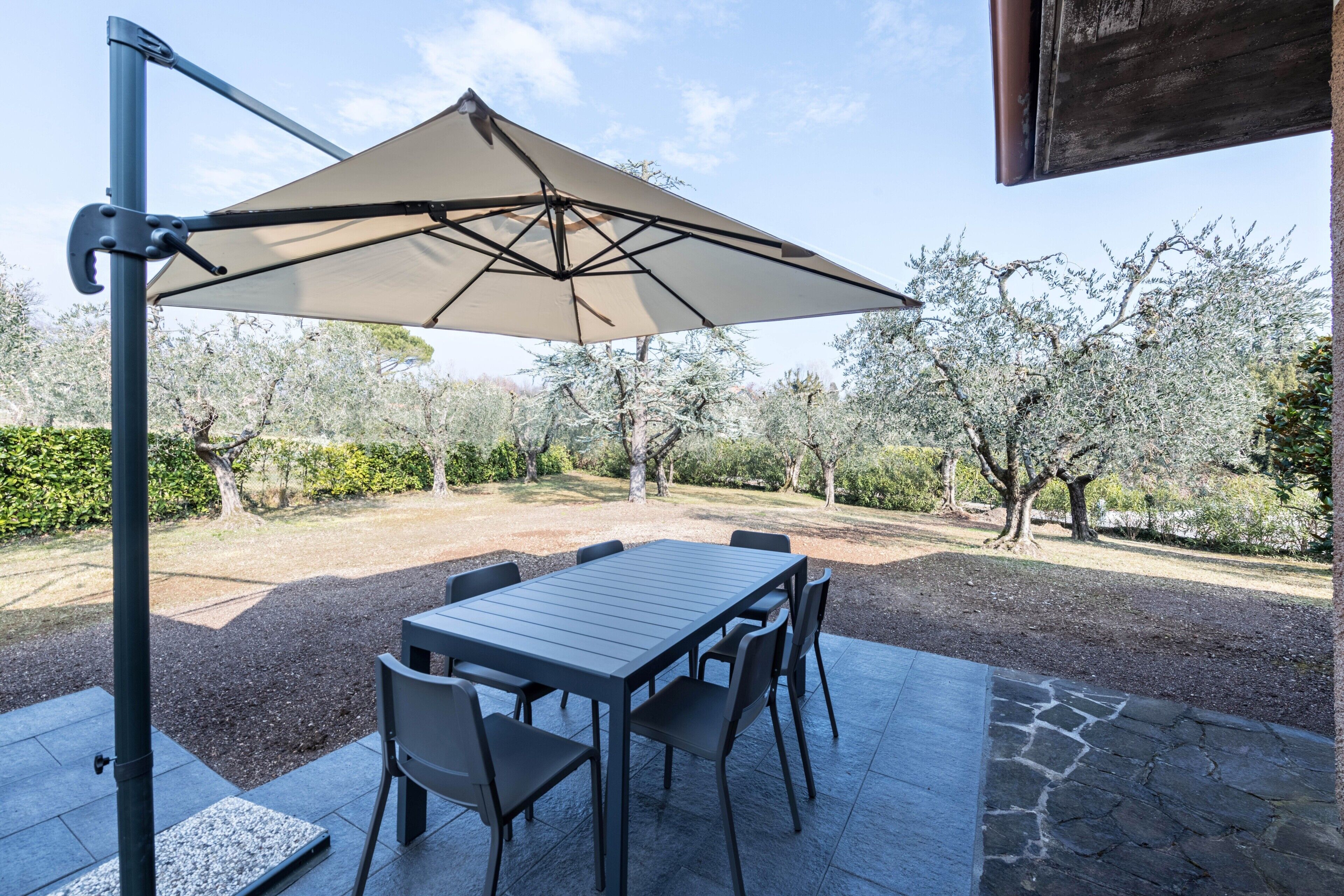 Un patio esterno con tavolo e sedie, un ombrellone grande e vista su alberi e un edificio.