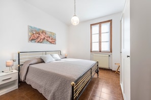 A bedroom with a bed, bedside table, lamp, painting, window, radiator, and a door.