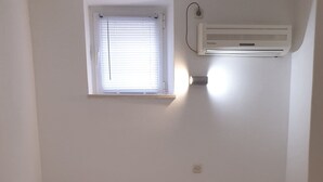 Ein Einzelbett mit Holz-Kopfteil, weißer Bettwäsche und einer gelben Decke. Ein Fenster mit Jalousien und eine Wand-Klimaanlage sind vorhanden.