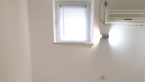 Ein Einzelbett mit Holz Kopfteil, weißer Bettwäsche und zwei Handtüchern. Ein Fenster mit Jalousien, eine Klimaanlage und eine Steckdose sind an der Wand angebracht.
