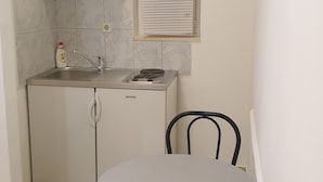 Eine kompakte Küche mit eingebautem Herd, Spüle und einem Fenster mit Jalousien. Ein kleiner Esstisch mit zwei Stühlen steht bereit.