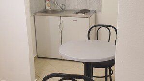 Eine kleine Küche mit weißen Schränken, einem Spülbecken und einem Fenster mit Jalousien. Ein runder Tisch mit zwei Stühlen steht daneben.