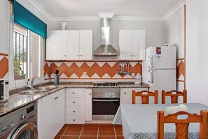 Une cuisine compacte avec des meubles blancs, une hotte de cuisinière en acier inoxydable, un four encastré, un lave-linge et un coin repas avec une table et des chaises.