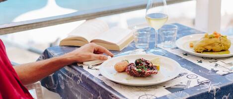 A person reading a book at a table with a nautical-themed tablecloth, a plate of food, and a glass of white wine.