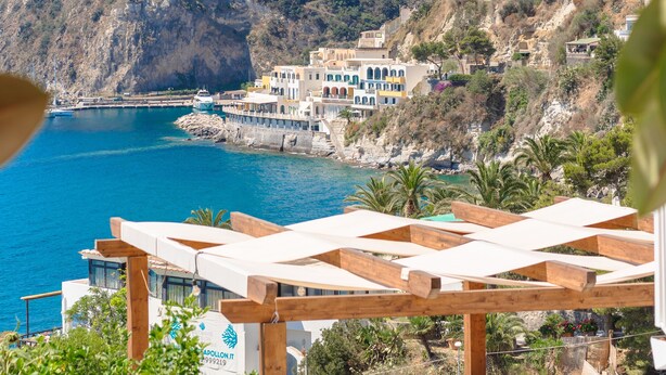 A coastal view with a wooden pergola, outdoor seating, and a building with a stone base. - Ferdinando Beach & Spa (Serrara Fontana)