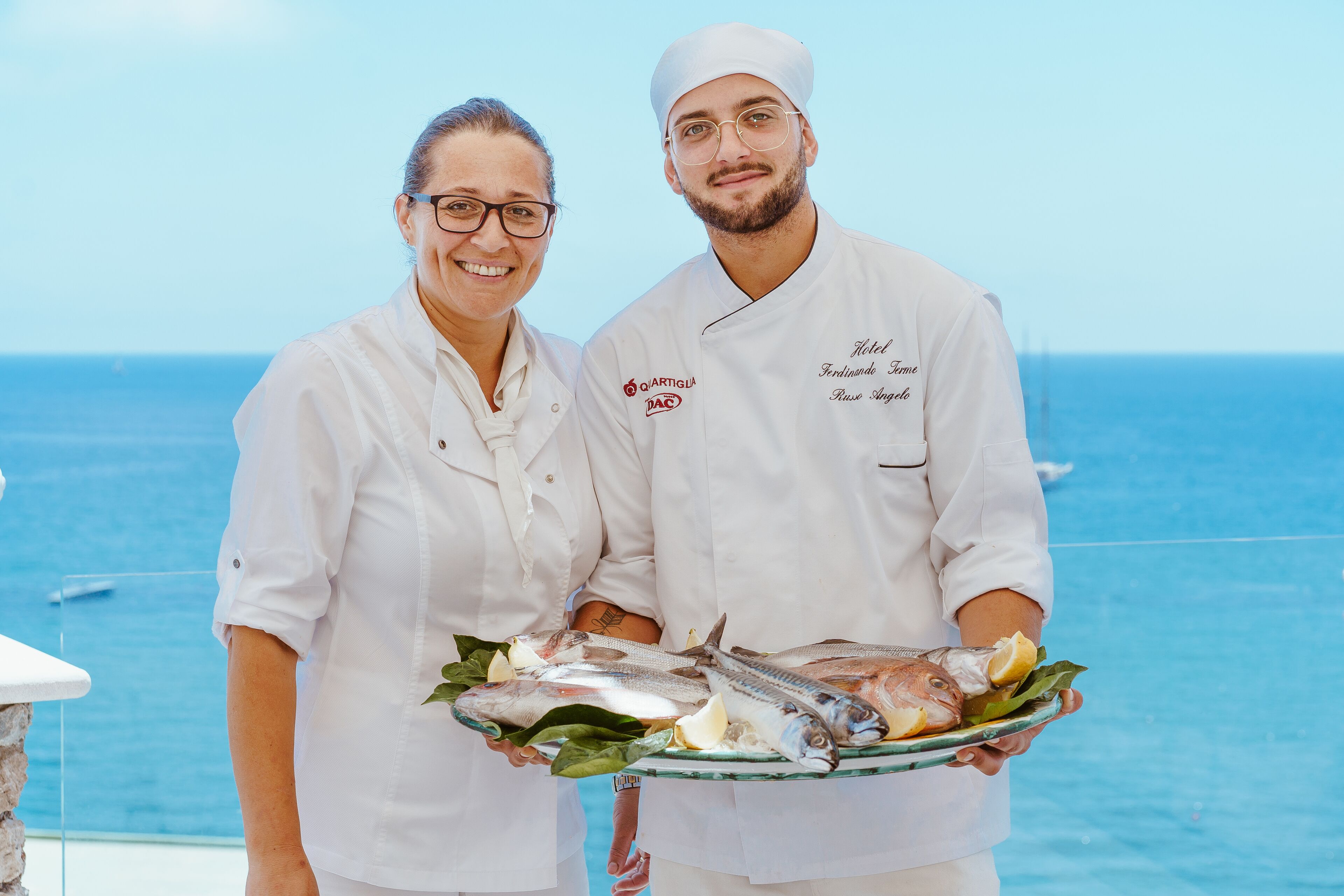 Two chefs in white uniforms holding a plate with fish and lemon slices, standing by a pool with a clear view of the ocean.