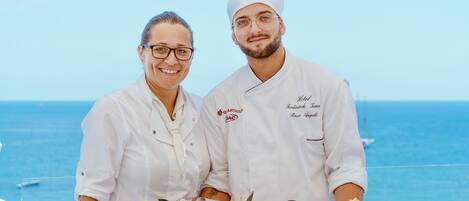 Two chefs in white uniforms holding a plate with fish and lemon slices, standing by a pool with a clear view of the ocean.