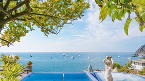 A pool with a view of the sea and boats, surrounded by greenery and a statue. - Ferdinando Beach & Spa (Serrara Fontana)