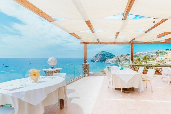 Un área de comedor al aire libre con mesas y sillas, con vista a la costa, barcos y montañas al fondo.