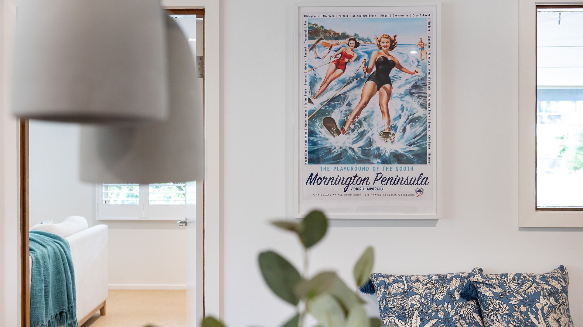 Une pièce décorée d’une affiche représentant la péninsule de Mornington, d’un canapé blanc et d’un coussin bleu à motifs.