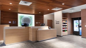 A hotel reception area with wooden reception desks, a large decorative wall piece, and a visible refrigerator stocked with beverages. - Fairfield Inn & Suites by Marriott Winnemucca (Winnemucca)