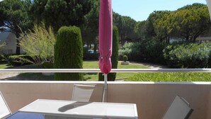 A balcony with a table and chairs, a pink umbrella, and a view of a well-maintained garden with various trees and shrubs. - Ground Floor Studio with Private Terrace and Parking in Port Camargue (Port Camargue)