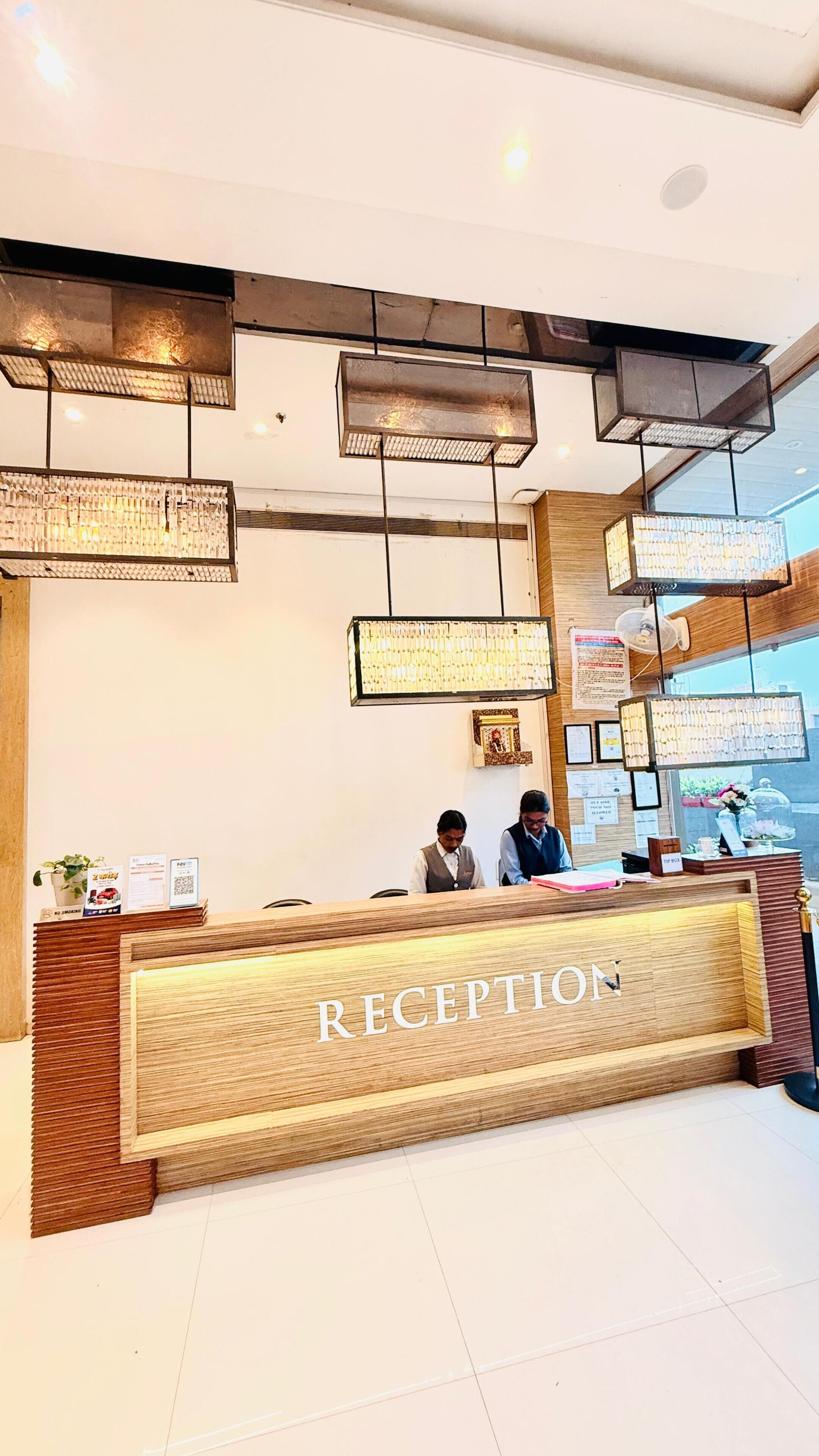 A reception desk with a wooden finish and the word 'RECEPTION' prominently displayed. There are two individuals seated behind the desk, and the area is well-lit with multiple hanging light fixtures.