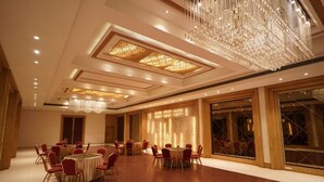 A spacious banquet hall with round tables and red chairs, a large chandelier, and a patterned wall feature. - Hotel Amora (Rajnandgaon)